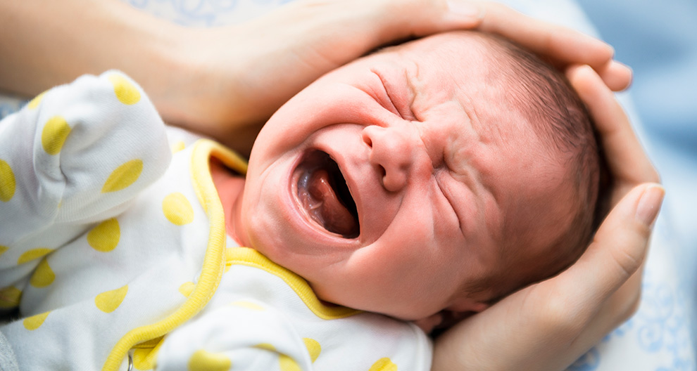 A close-up of a crying baby's face, held by a hand, with a white and yellow polka-dotted outfit.