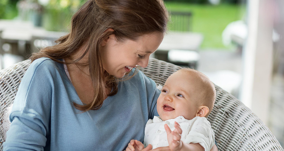 A woman in a blue shirt is holding a baby in a white outfit, both looking at each other, with a blurred background of a patio setting.