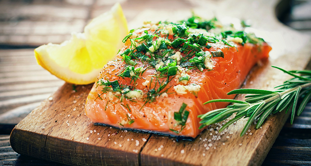 A salmon fillet seasoned with herbs and garlic, accompanied by a lemon wedge and rosemary sprig, presented on a wooden cutting board.
