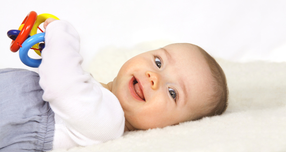 A baby lies on a white surface, holding a colorful toy.