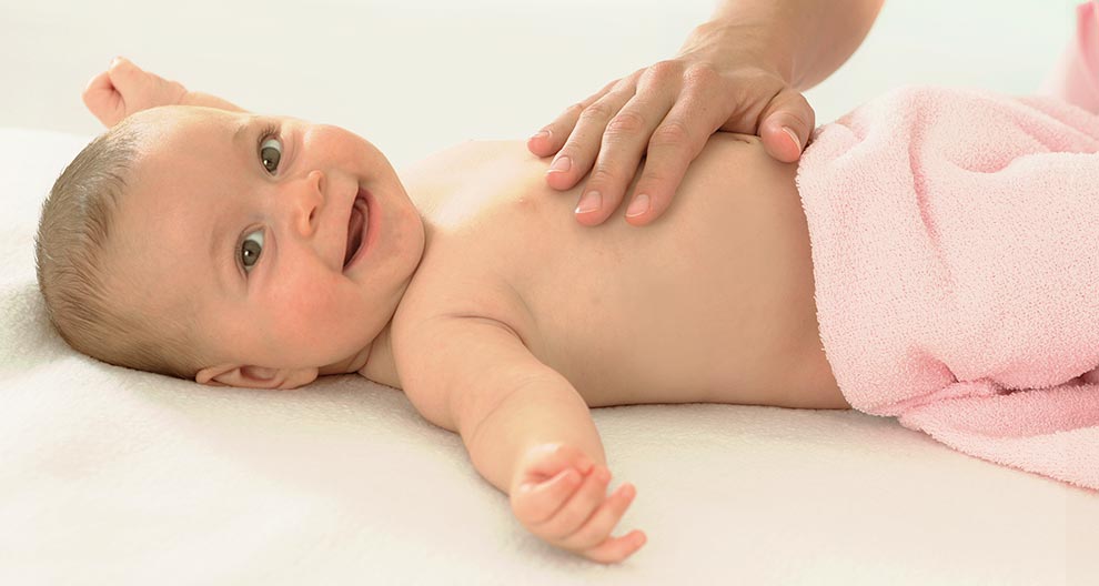 A baby lies on a white surface, wrapped in a pink towel, with a hand placed on the baby's chest.