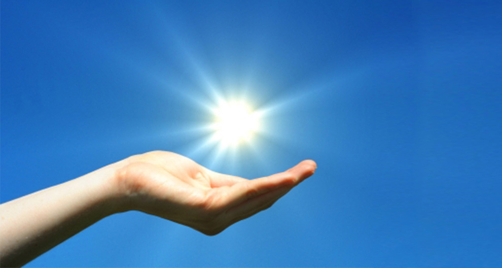 A hand holds a radiant sun against a clear blue sky.
