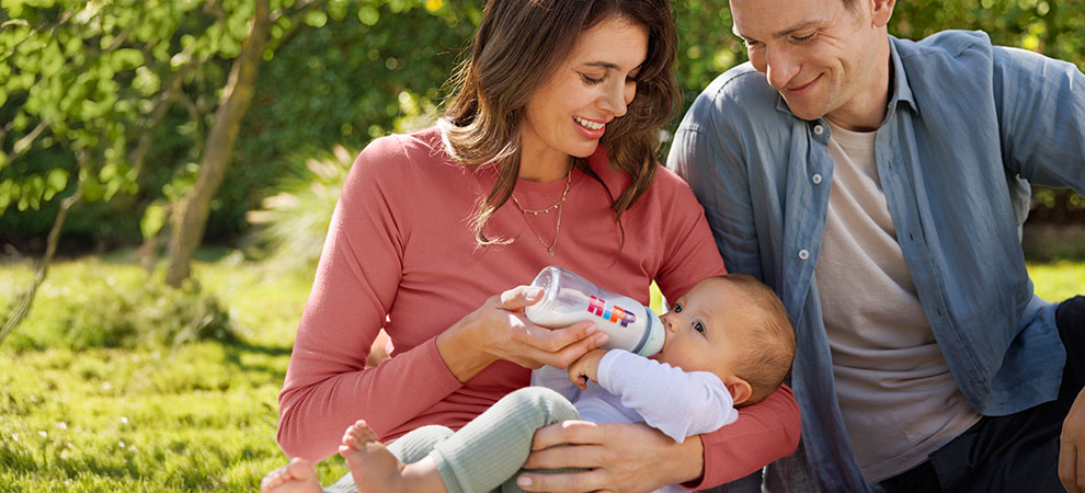 A man and a woman sit outdoors, feeding a baby with a bottle.